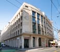 A large, multi-story commercial building with a distinctive Art Deco-style facade. The building has a prominent sign that reads 'RHONE' and features decorative elements like carved stone figures. The building is located on a busy city street with tram lin