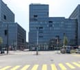 Three high-rise buildings with glass facades, street with crosswalks, parked cars, trees, bus stop, street signs, and a tram in the distance.