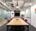 Conference room with a long wooden table, surrounded by red and gray chairs. The room has glass walls and a glass wall to the left leading to another room. A TV is mounted on the wall, and there is a clock above the shelf with a potted plant on it. The ro