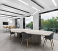 Large open office space with a long wooden conference table, gray upholstered chairs, and a large window overlooking a wooded area. The ceiling has recessed lighting fixtures and the floor is a light-colored wood laminate. There is a TV mounted on the wal