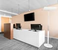 Modern office space with white reception desk, computer monitors, and a large TV mounted on the wall. The room has beige walls, a gray striped floor, and recessed lighting fixtures.