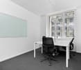 The image shows a modern office space with a large white board on the wall, a desk with a computer and office chair, and large windows overlooking a city skyline. The room has a minimalist and professional design with white walls, gray carpeting, and a cl
