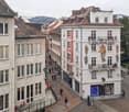 This image shows a picturesque street in a European city, with a mix of historic and modern buildings. The street is lined with shops and cafes, and there are people walking along the sidewalks. The buildings have ornate architectural details, including b
