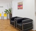The image shows a modern and minimalist living room. The room has hardwood floors, white walls, and a black leather sofa set. There is a decorative plant and artwork on the wall. The room also features a shelving unit with yellow and gray accents.