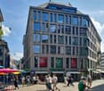 modern building with many windows, stores on the bottom floor, people walking on the street, umbrellas on the side