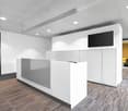 Modern, minimalist office space with white walls, recessed lighting, a reception desk, and a striped carpet floor. There is a TV mounted on the wall and what appears to be a dishwasher or other appliance behind the reception desk.