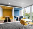 This image shows a modern and colorful office space with large windows providing a view of the surrounding greenery. The space features a yellow and blue color scheme, with yellow chairs and a yellow seating area, as well as a blue seating area. There is