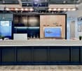 The image shows a modern and stylish office space for the company Regus. The space features a large reception desk with a concrete countertop, black cabinets, and a display screen showing the Regus logo. The walls are decorated with framed artwork and map