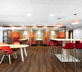 Modern and colorful office space with wooden flooring, white tables, red chairs, and patterned wall panels. The space includes a dishwasher and appears to be designed for collaborative work or a casual office environment.