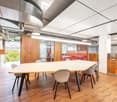 Large open office space with wooden walls, hardwood floors, and a long conference table with chairs. The ceiling has exposed ductwork and lighting fixtures. There are also storage cabinets and a dishwasher visible in the space.