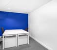 Small office space with a white table, two black chairs, and a bright blue accent wall. The room has a white ceiling and gray carpeted floor.
