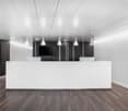 Large open office space with white reception desk, black accent walls, and multiple pendant lights hanging from the ceiling. The floor is made of dark hardwood. There is a small kitchenette area visible in the background.