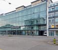 A large glass and steel commercial building with the Regus and Buche logos. The building has multiple floors and entrances, with a parking area in front. There are people walking around the building and a car parked in the parking area.