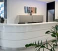 The image shows a modern and stylish office reception area. The reception desk is a curved, white counter with a sleek design. Behind the desk, there is a large abstract artwork on the wall and a cabinet with a decorative vase on top. The room has large w