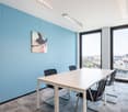 Spacious meeting room with large windows offering a view, light-colored table, black chairs, and a modern artwork on the blue wall