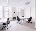 Spacious office space with large windows, white walls, and modern furniture. The room features a long table with several black office chairs, as well as individual desks and chairs. The space appears to be well-lit and designed for a professional work env