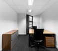 Small office space with white walls, black and wood furniture, including a desk, cabinet, and office chair. The room has a window and a suspended ceiling with lighting fixtures.
