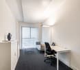 The image shows a modern office space with a large window, a desk with a computer, a chair, and a sofa. The room has white walls and a gray carpet floor. There is also a cabinet and a decorative object on a side table.