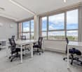 Spacious office space with large windows providing a scenic view, white desks and black office chairs, modern lighting fixtures, and a carpeted floor.