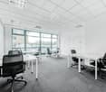 An empty office space with white walls, carpeted floor, large glass windows, and multiple white desks with black chairs.