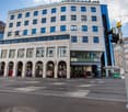 Multi-story commercial building with a modern, concrete facade and large windows. The ground floor has several retail stores with display windows. The upper floors appear to be office or residential spaces. The building is located on a street with tram li
