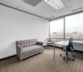 The image shows a small office space with a gray sofa, a desk with a chair, and large windows providing natural light. The room has a neutral color scheme with white walls, gray carpet, and a suspended ceiling with recessed lighting.