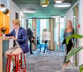 Modern and colorful office space with open layout, high ceilings, and large windows. The space features a kitchen area with a dishwasher, as well as various seating and work areas. The decor includes plants, bright lighting fixtures, and a mix of furnitur