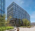 A modern, multi-story office building with a glass facade and wooden accents. The building is surrounded by lush greenery, including trees and landscaped areas. There is a paved plaza in front of the building with seating areas.