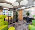 Modern and open office space with large windows, green and yellow upholstered furniture, glass meeting pods, and decorative plants