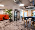 Open office layout with modern furniture, including orange sofas, a large conference table, and bar stools. The space features high ceilings, pendant lighting, and large windows. There are also several potted plants throughout the room, adding a touch of