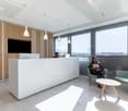 Modern office space with large windows providing a view, white reception desk, wooden accent wall, pendant lighting, and comfortable seating area with green chairs and a coffee table.