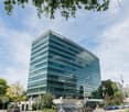 A modern, multi-story office building with a glass facade and the Deloitte logo prominently displayed. The building is surrounded by trees and has a parking area in front.