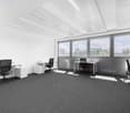 Large open office space with white walls, gray carpeted floor, multiple desks and office chairs, and large windows providing natural light and a view of the outdoors.