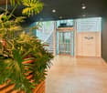 Open entrance hall with potted plants, clean and minimalist, white elevator doors, light wood floor, dark walls, logo of Regus on the wall