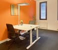 The image shows a modern office space with an orange accent wall, a white desk with a height-adjustable base, a black office chair, and a potted plant. The room also has a large window providing natural light and a carpeted floor.