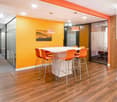 Bright and modern office space with orange and yellow color scheme, large white table with orange bar stools, wood-look flooring, and glass walls separating different areas