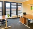 The image shows a modern office space with large windows, yellow walls, and wooden furniture. The room has two desks with chairs, a green vase on one of the desks, and a wall-mounted artwork. The floor is covered with gray carpeting.