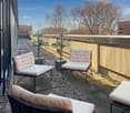 Outdoor patio with comfortable seating, including two armchairs and a sofa, surrounded by potted plants and a concrete wall. The patio has a gravel floor and is accessible through large windows or doors.