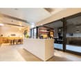 Open-plan office space with a reception desk, dining area with tables and chairs, and large windows providing natural light. The space has a modern, minimalist design with white walls and floors.