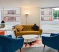 Contemporary room with large windows, yellow couch, colorful rug, modern chairs, and two white coffee tables.