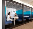 Conference room booth with blue upholstered benches, white table, grey and blue partition walls, pendant lights, and a drawing on the wall.