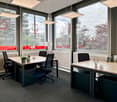 An office room with a large windows overlooking the trees and buildings, multiple desks with office chairs, pendant lamps, a lamp on the desk, some potted plants and lamps on the desks.