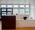 The image shows a modern office space with a reception area. The reception desk is made of dark wood and has a computer monitor and other office supplies on it. The office has large windows that provide natural light and a view of the outdoors. The ceilin