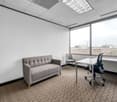 This image shows a modern office space with a gray sofa, a desk with a chair, and large windows providing a view of the exterior. The room has a suspended ceiling with recessed lighting and a carpeted floor.