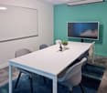 A meeting room with a long white table, six chairs, a whiteboard, a TV, and a potted plant on the table.