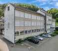 Multi-story office building, modern architecture, multiple cars parked, concrete parking lot, green surroundings