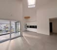 Bright, empty room with white walls and ceiling, large grey tiles on the floor, skylight, sliding glass doors to a balcony, white kitchen with modern appliances, white cabinets.