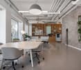 Spacious open-plan office with modern furniture, including a large conference table, swivel chairs, and a built-in bookshelf. The space features high ceilings, exposed ductwork, and large windows providing natural light. There is also a potted plant and a