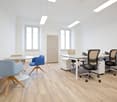 Bright and modern office space with large windows, white walls, hardwood floors, a round table with chairs, a desk with office chairs, and a storage cabinet.