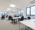Large open office space with multiple workstations, desks, and chairs. The room has large windows providing natural light and a view of the city outside. The ceiling is equipped with recessed lighting fixtures. The overall design appears modern and minima